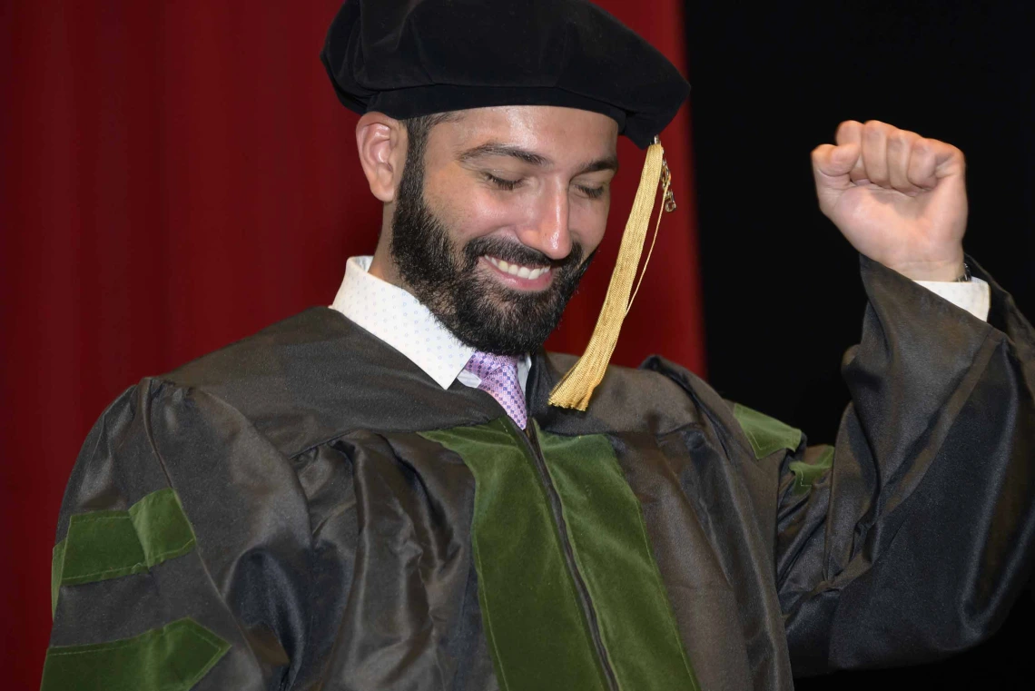 A medical student wearing graduation regalia pumps his fist and smiles onstage during a graduation ceremony. 