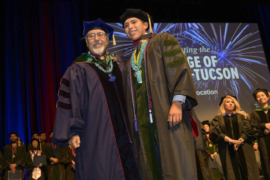 A professor and medical student in graduation regalia stand together onstage during a graduation ceremony. 