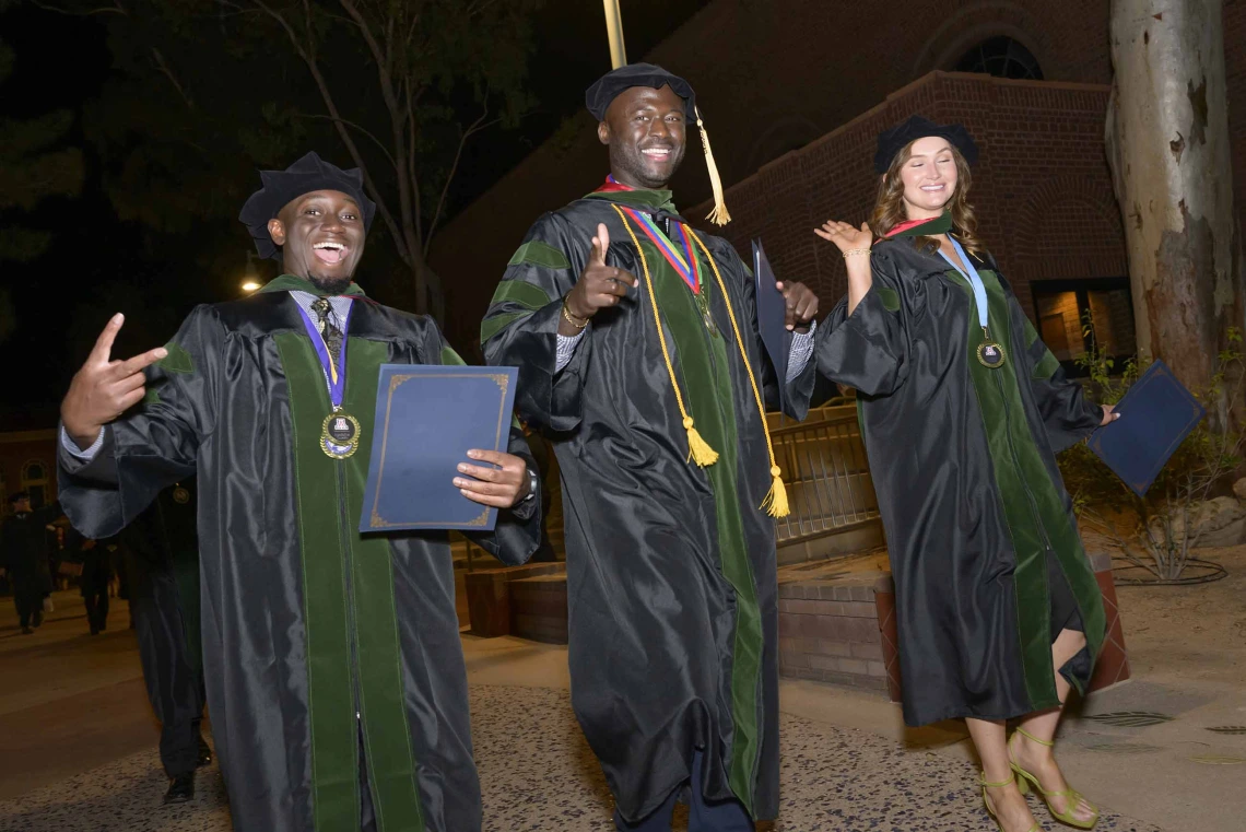 Three medical students in graduation regalia wave in celebration while walking outside at night. 