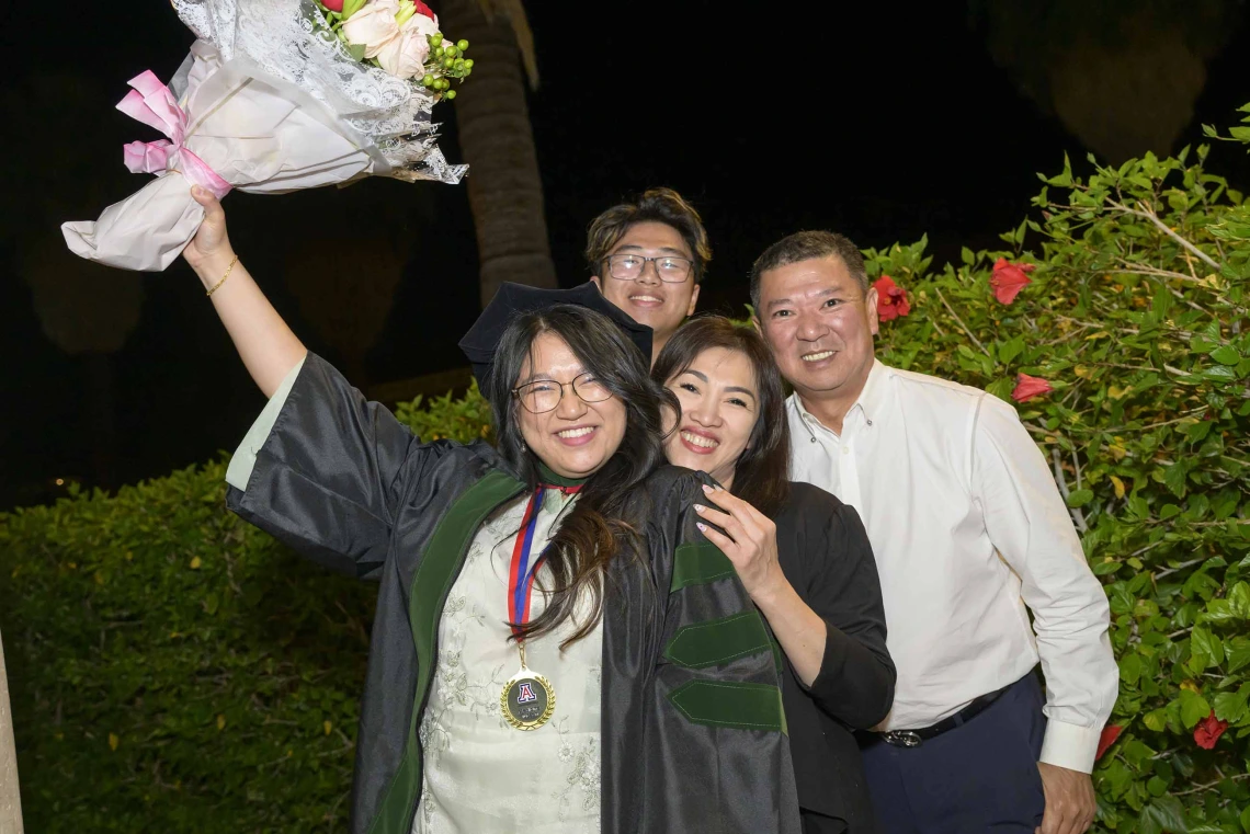 A medical student in a graduation robe holds flowers up in celebration as her smiling family stands behind her. 