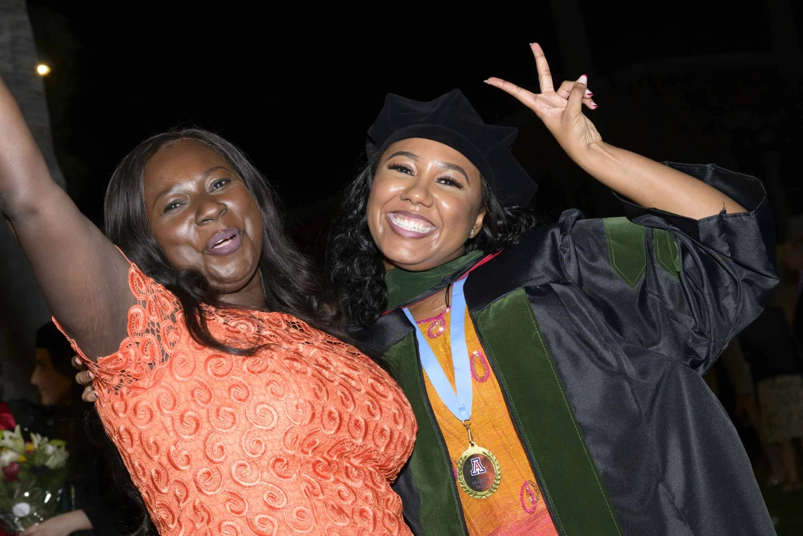 A medical school graduate dressed in a cap and gown, hugs her mother as they raise their hands in the air in celebration. 