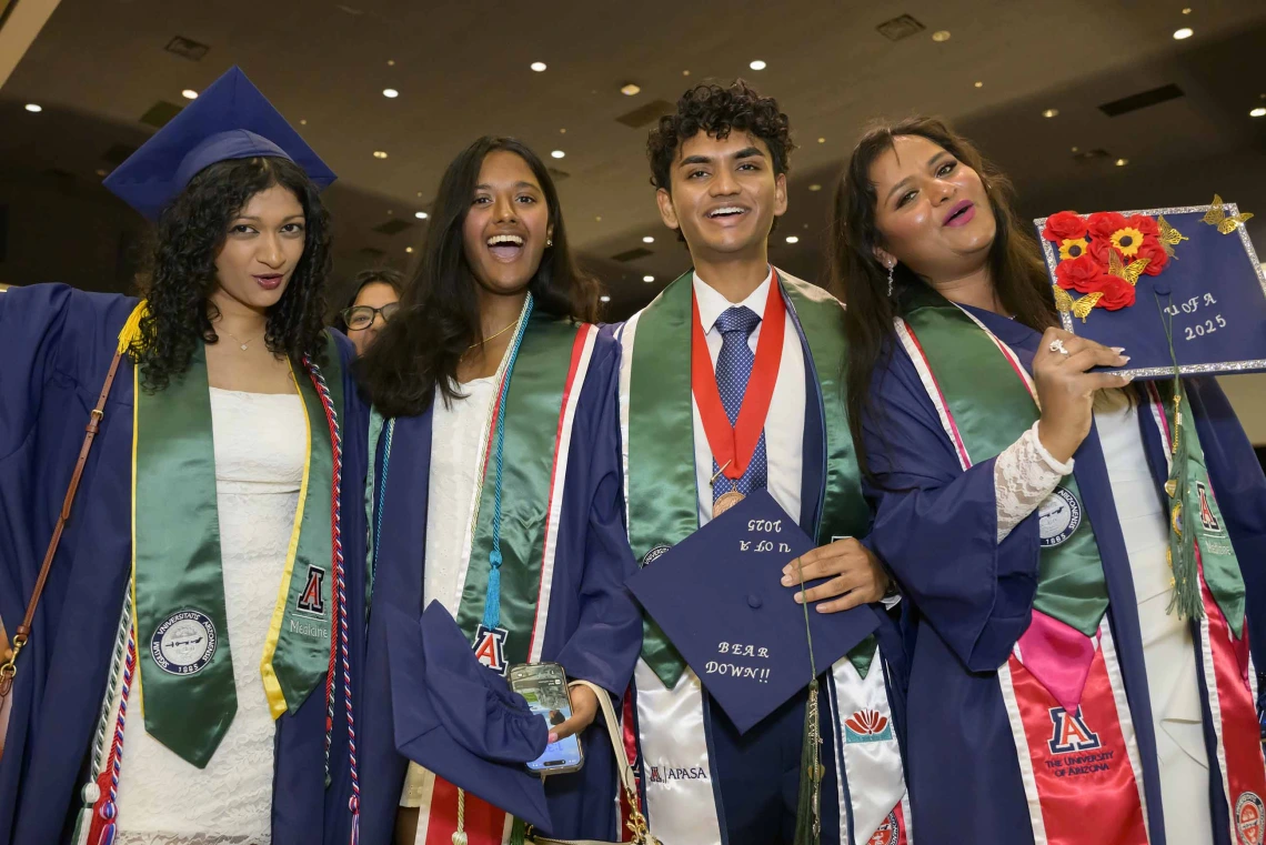 Four college students dressed in caps and gowns smile and cheer at a graduation ceremony. 