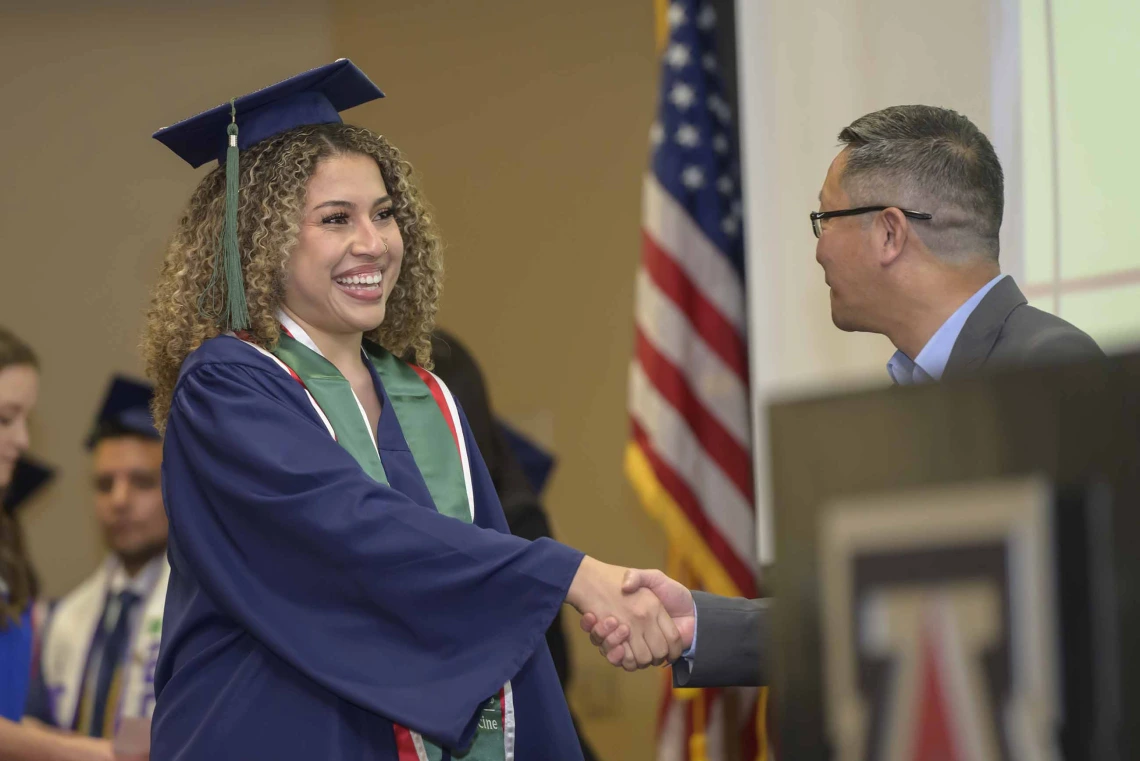 A young woman in a graduation cap and gown smiles as she shakes hands with a man during a graduation ceremony. 