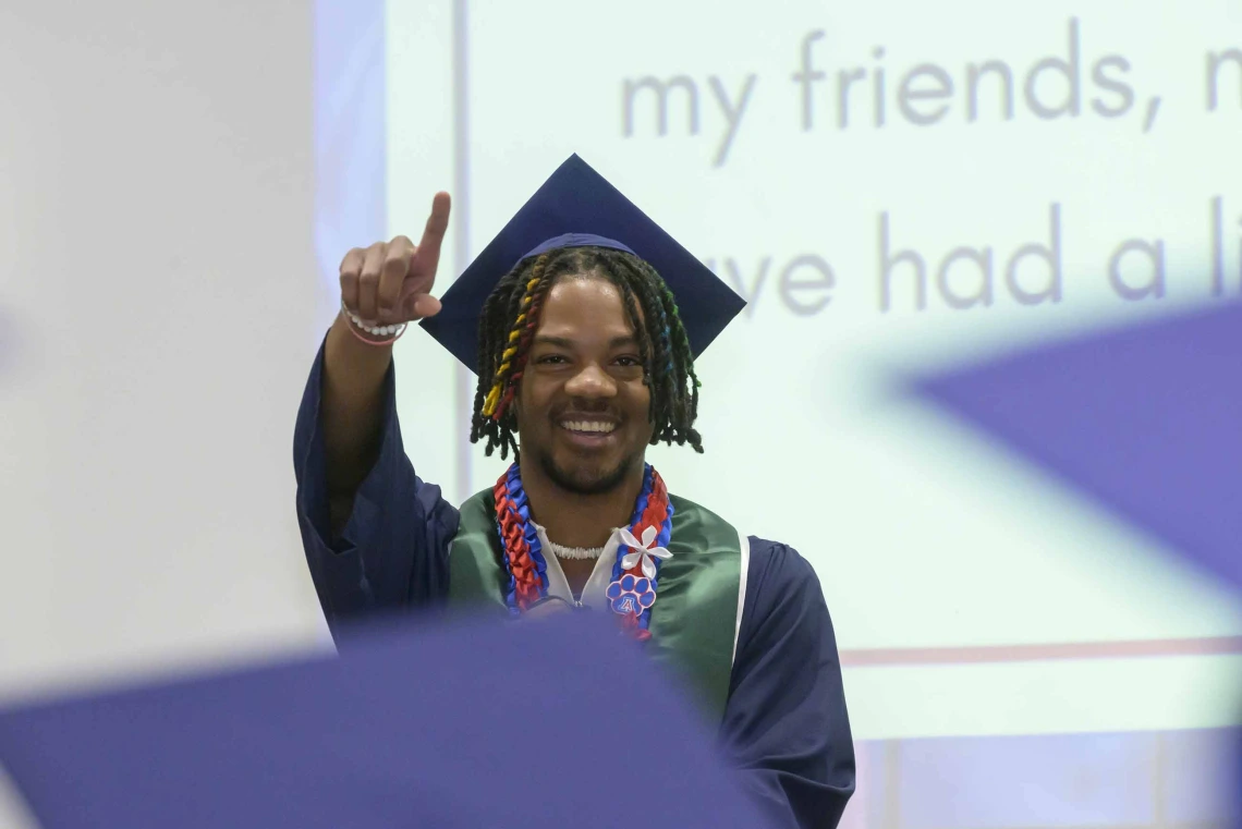A student wearing graduation regalia stands onstage smiling and holding up a finger in celebration. 