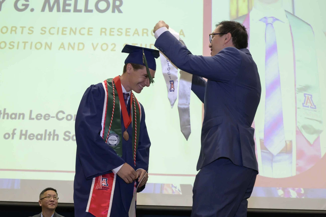 A young man wearing a graduation cap and gown is presented with a white sash onstage by a professor during a graduation ceremony. 