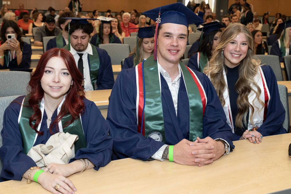 Several students in graduation caps and gowns sit in a room, smiling. 