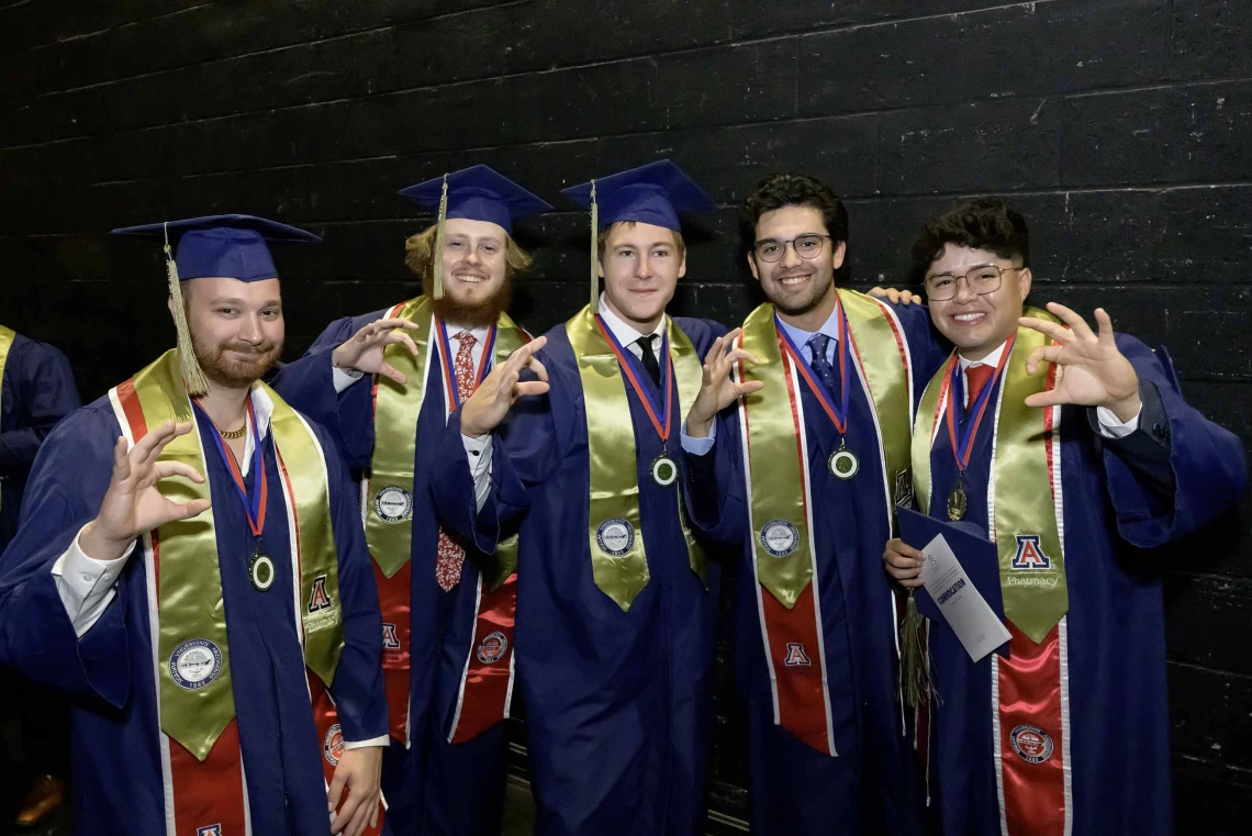 Five young men wearing graduation caps and gowns stand together, smiling and making the hand sign for the Arizona Wildcats. 