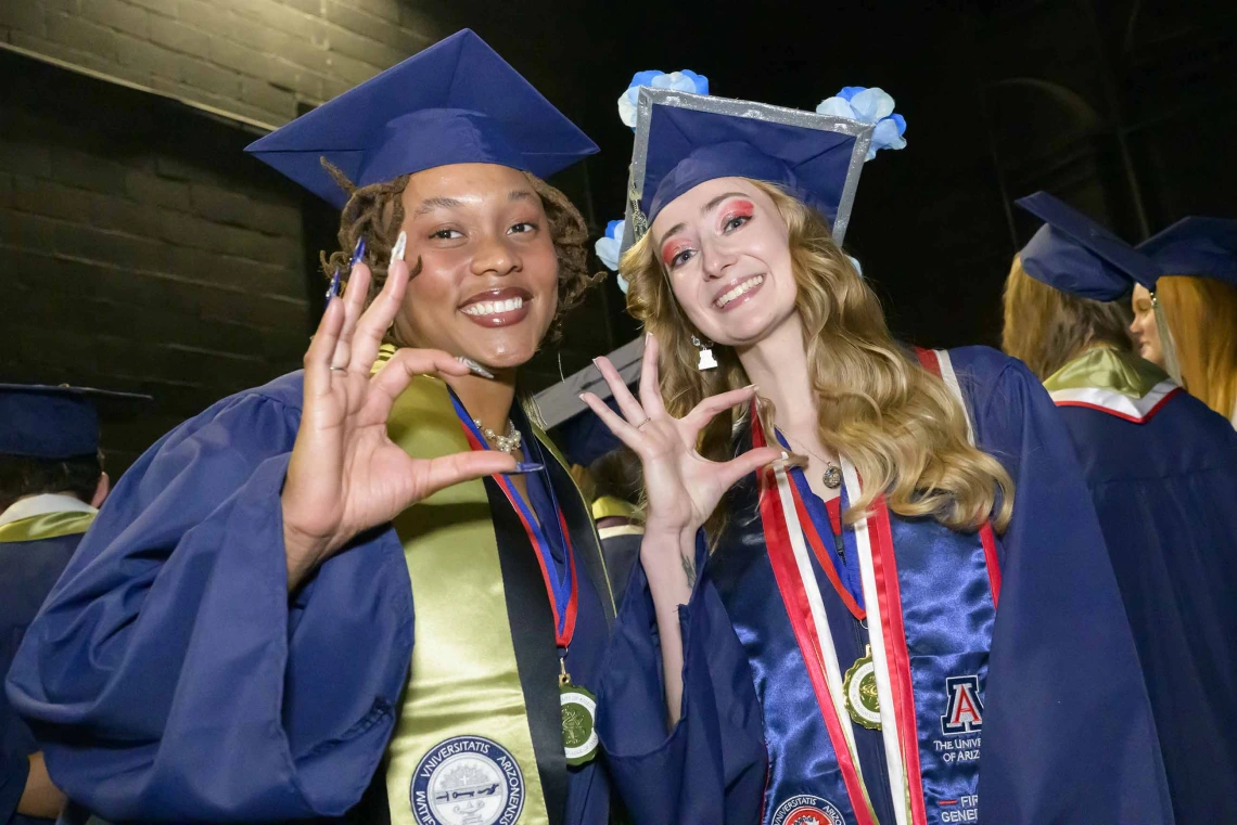 Two young women in graduation caps and gowns smile while giving the hand sign for the Arizona Wildcats. 