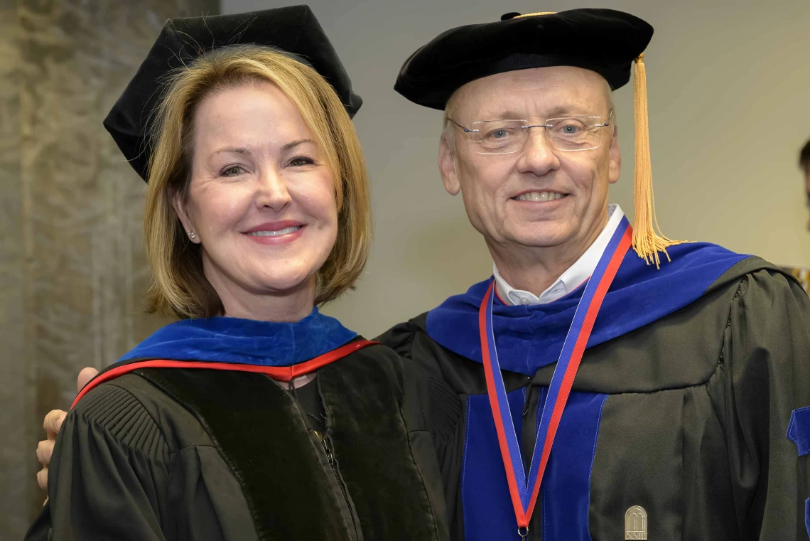 Two professors in graduation regalia stand together, smiling. 