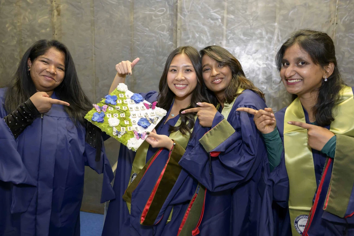 Four young women dressed in graduation regalia smile as they hold up a decorated graduation cap. 