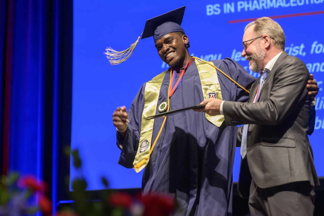 A young man dressed in a graduation cap and gown smiles as he is handed his diploma by an older man in a suit. 