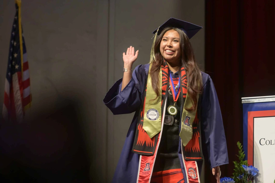 A young woman wearing graduation regalia smiles and waves as she walks onto a stage. 