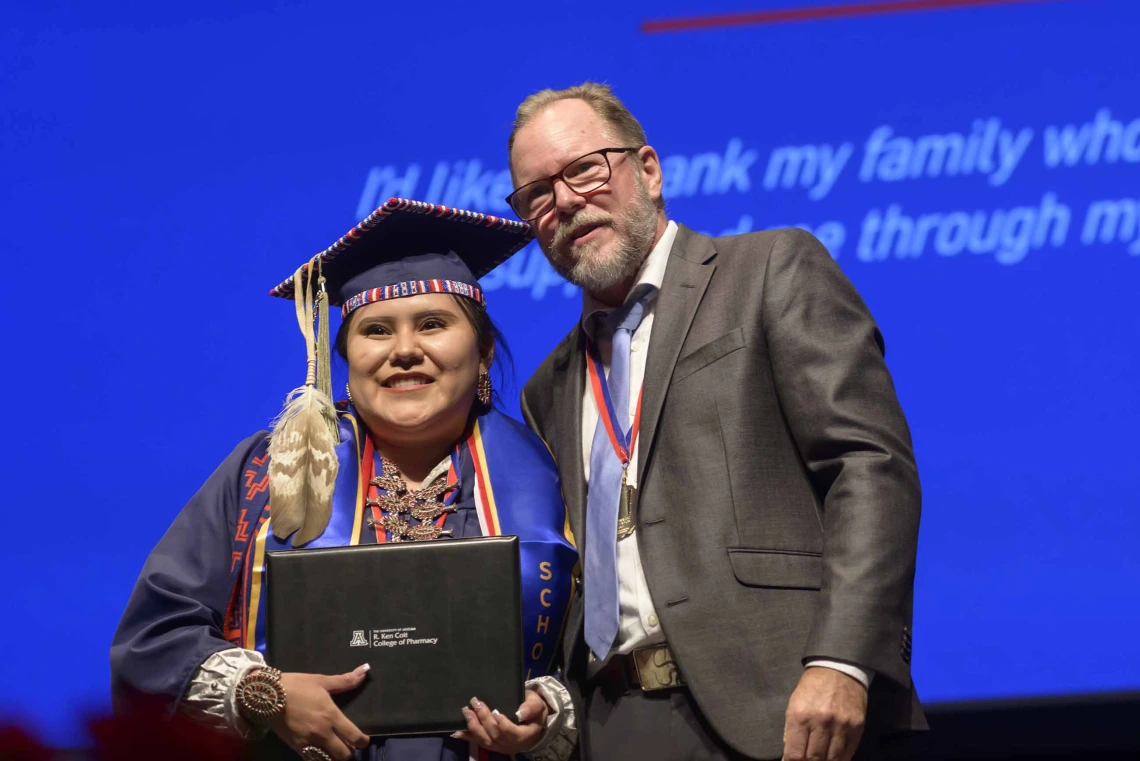 A young woman dressed in a graduation cap and gown holds her diploma and smiles as a professor in a suit stands next to her. 