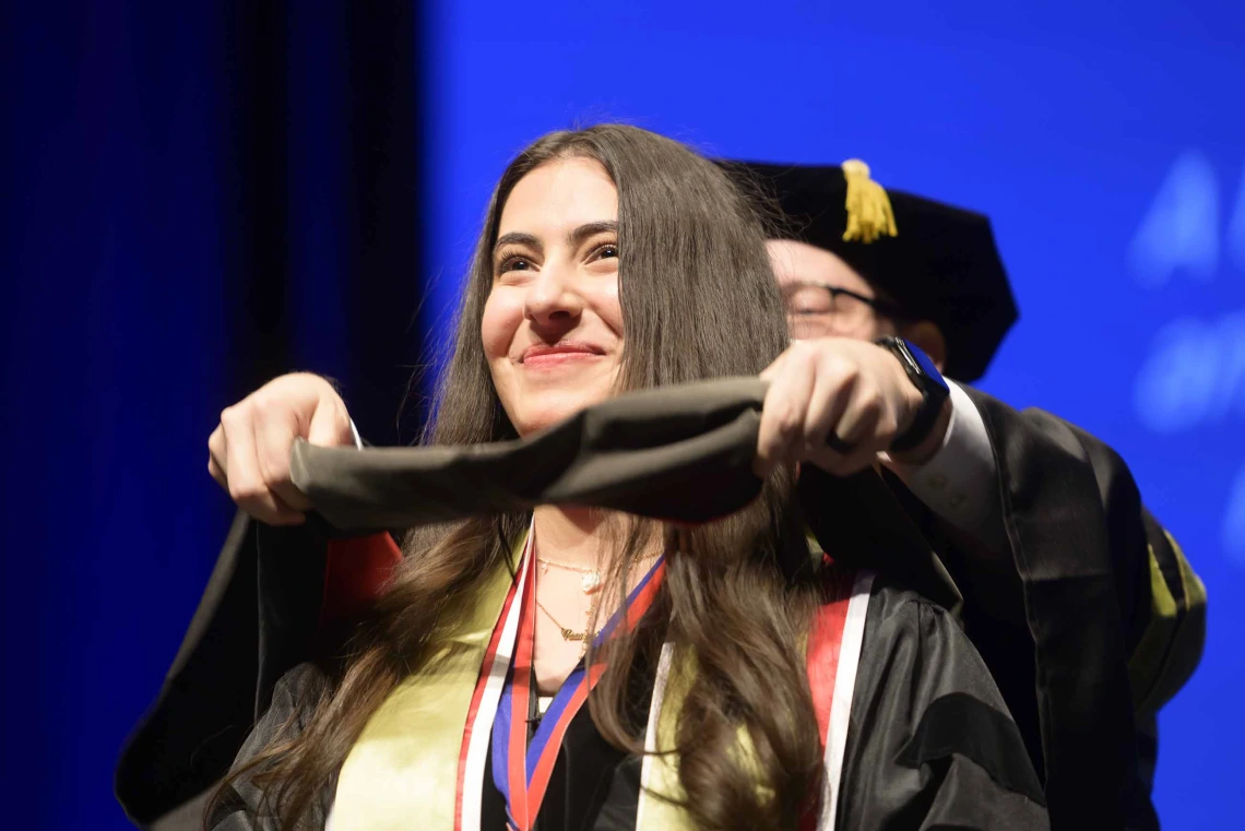 A young woman dressed in a graduation gown smiles as a professor places a ceremonial hood over her shoulders. 