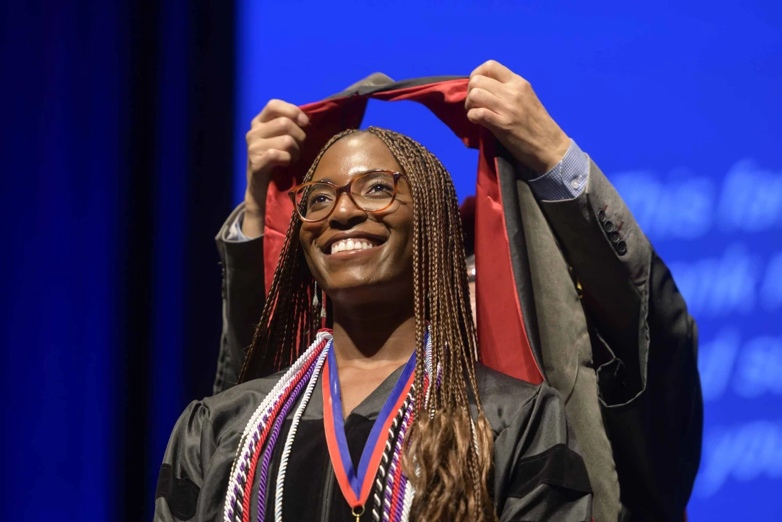 A young woman dressed in a graduation gown smiles as a professor places a ceremonial hood over her shoulders.