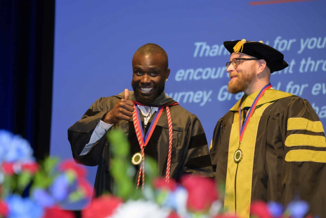 A young man in graduation regalia smiles and gives a thumbs-up while standing next to a professor who is wearing a cap and gown. 