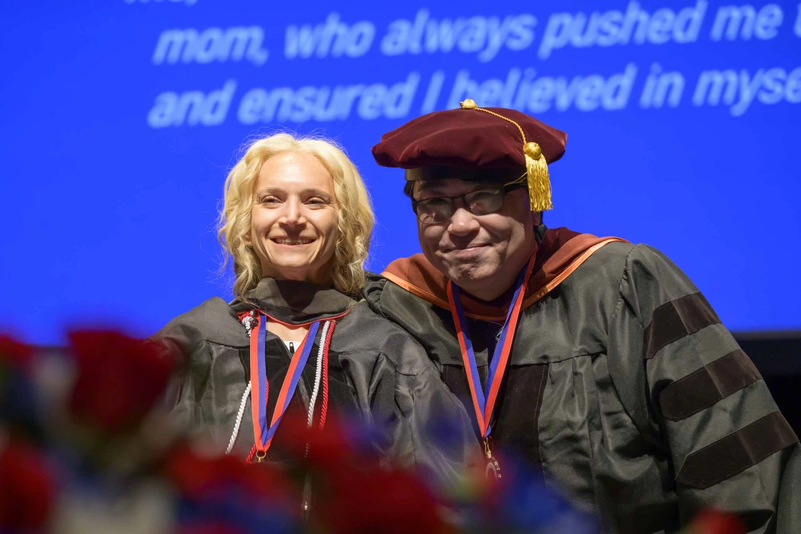 A young woman dressed in a graduation gown smiles as a professor, also dressed in graduation regalia, smiles behind her. 