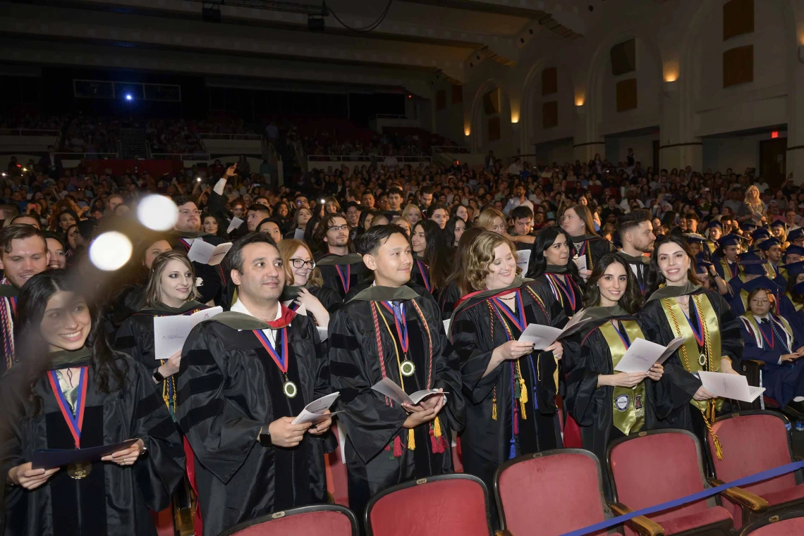 More than 225 students dressed in graduation regalia stand in an auditorium at the conclusion of their convocation ceremony. 