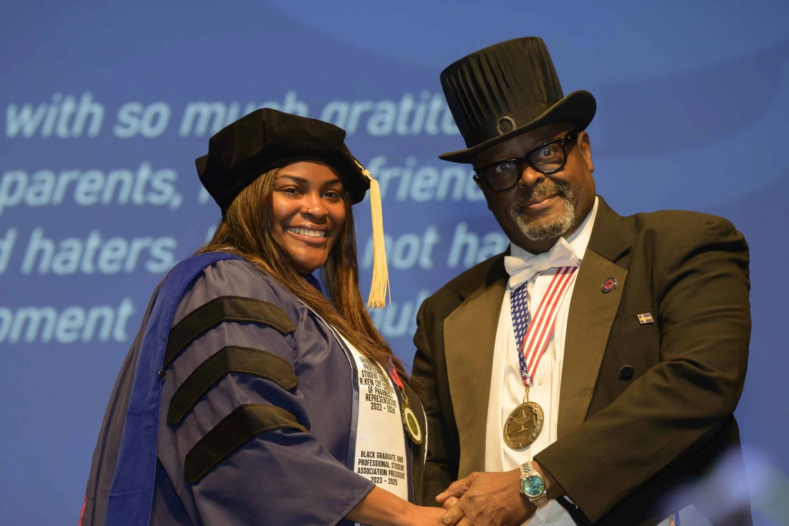 A student in a cap and gown shakes hands with a professor in a suit and top hat during a graduation ceremony.