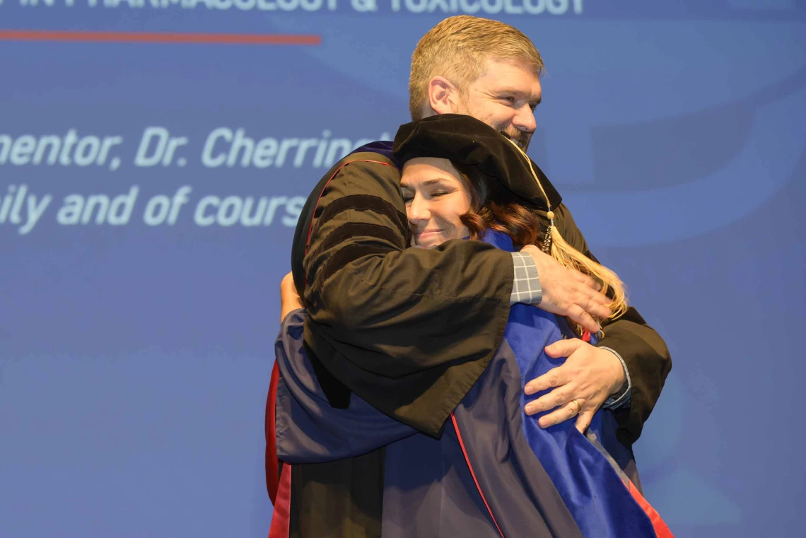 A professor hugs a woman onstage during a graduation ceremony. Both are dressed in graduation regalia. 