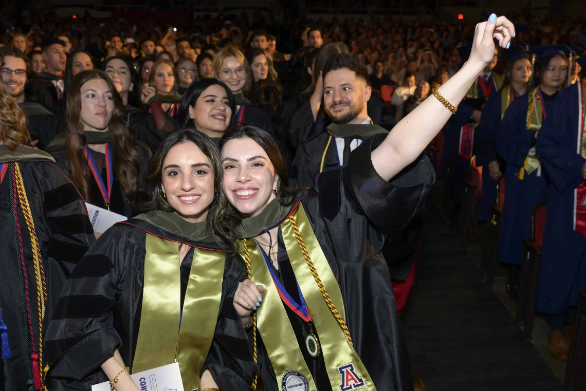 Two women dressed in graduation regalia smile while one raises her arm in celebration. They are standing in front of dozens of other graduates. 