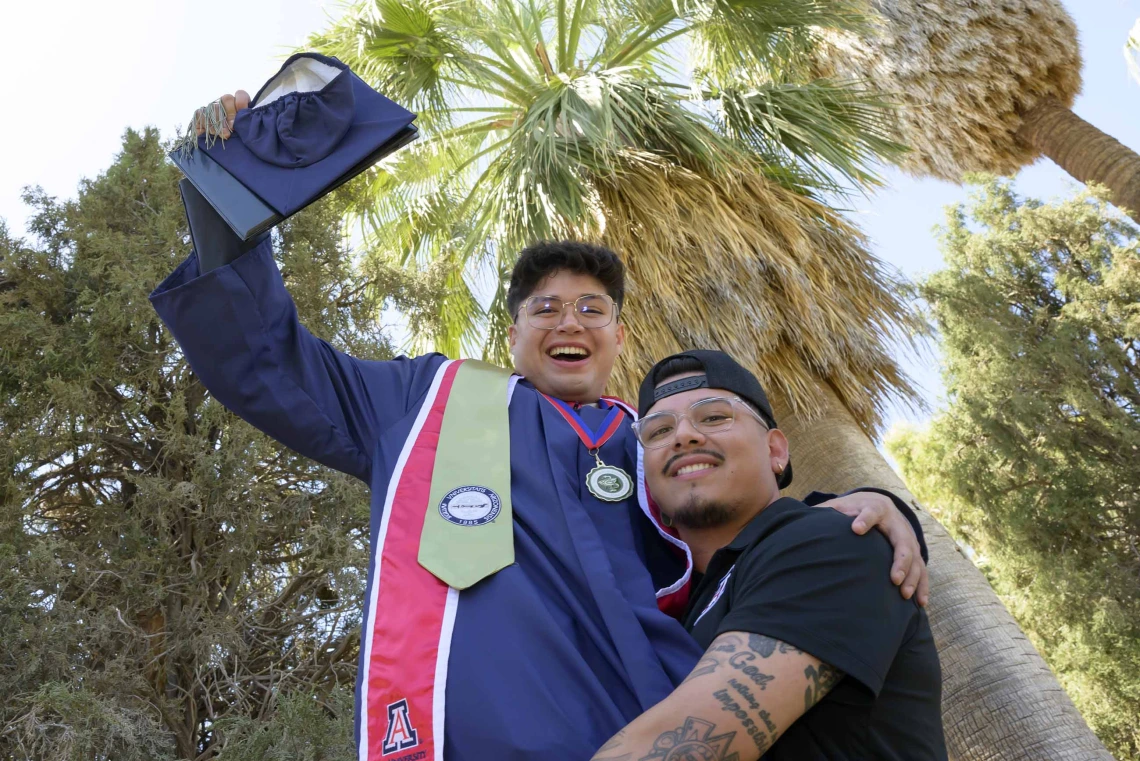 A young man in graduation regalia is hoisted into the air by another young man in celebration. Both are smiling outside under some trees.