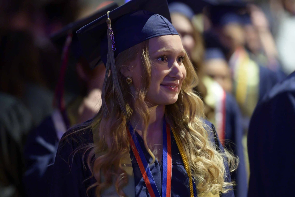 A young woman in a graduation cap and gown smiles as she looks off into the distance with other graduates behind her. 