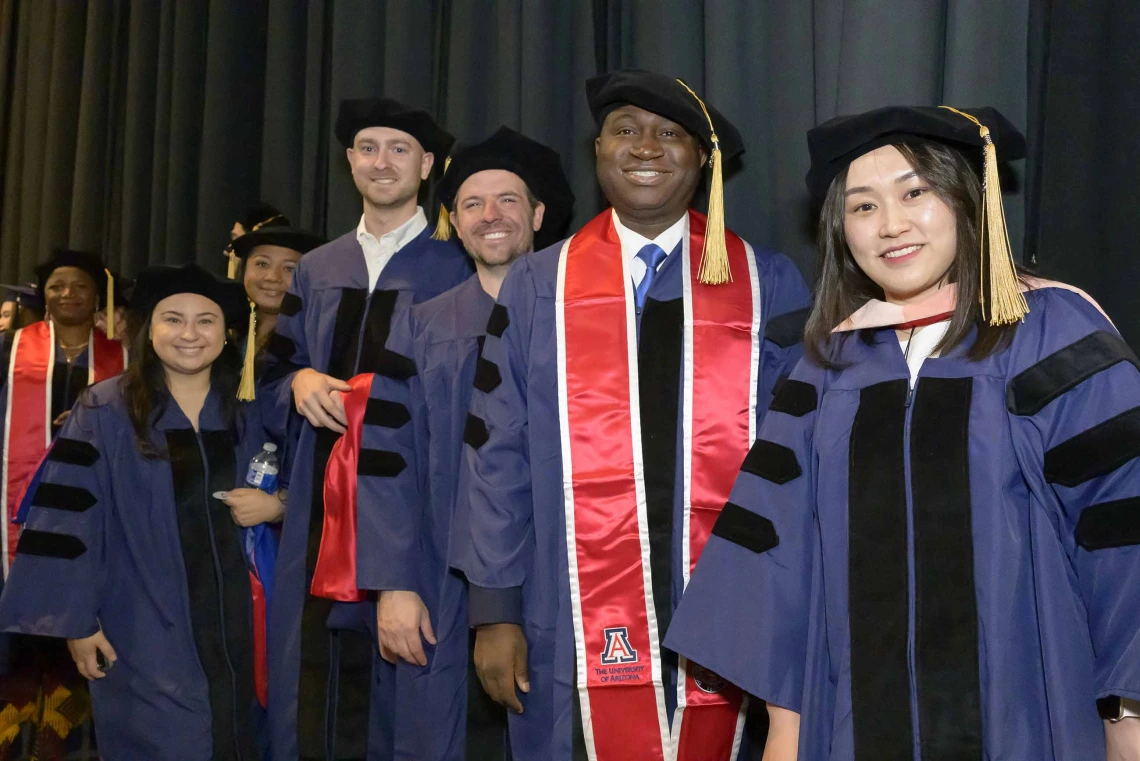 About a dozen doctoral students wearing graduation regalia smile as they stand in line in front of dark stage curtains. 