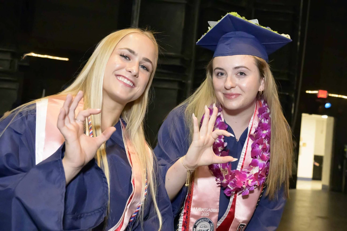 Two young women in graduation regalia smile and give the Arizona Wildcats hand sign.