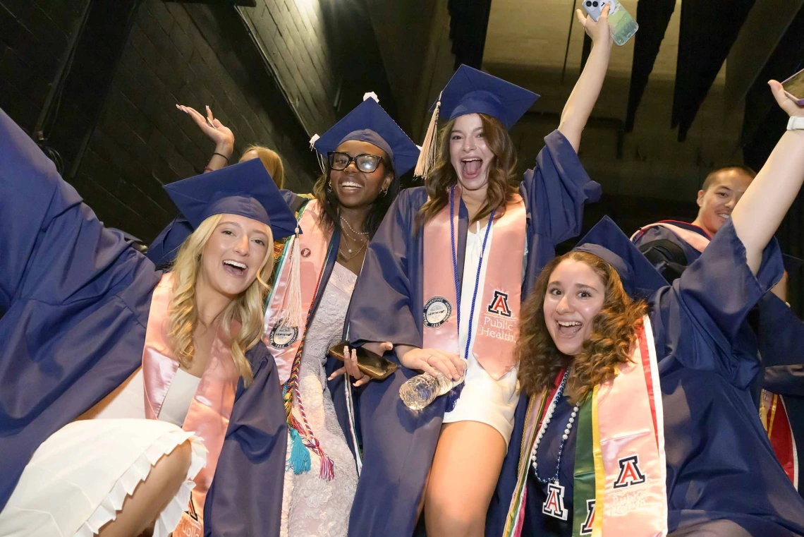 Four young women in graduation caps and gowns raise their hands and cheer in celebration. 