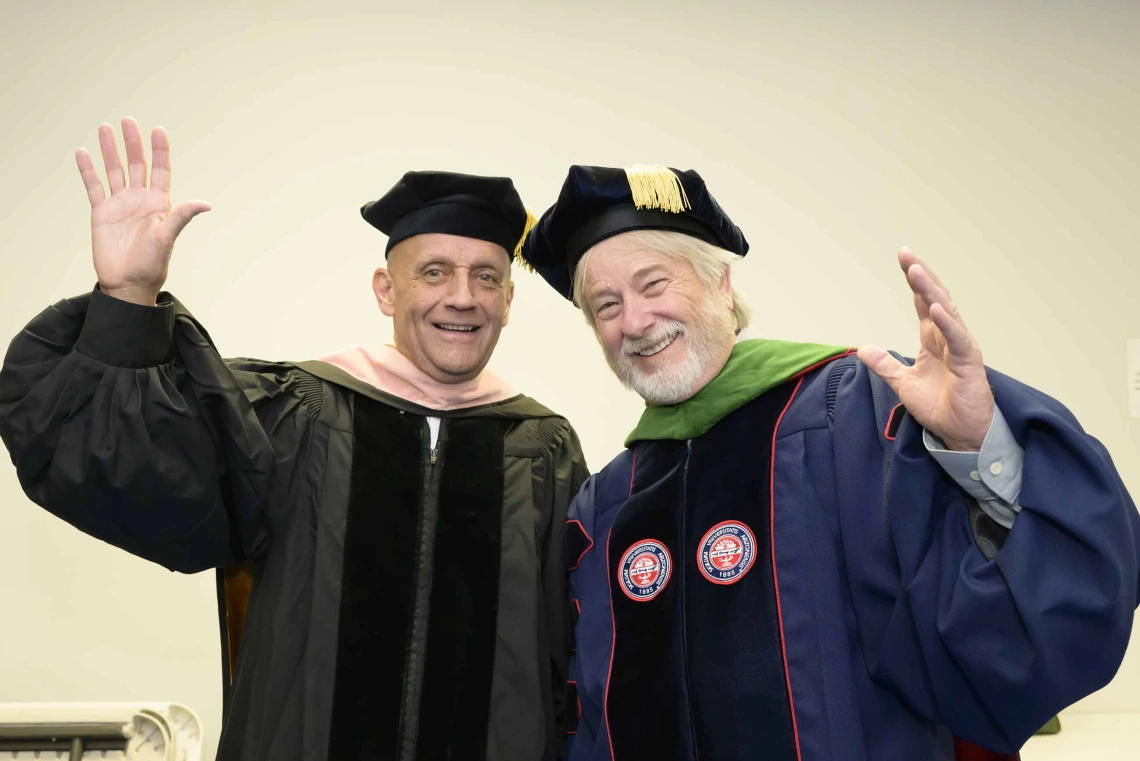 Two professors in graduation regalia smile and wave. 