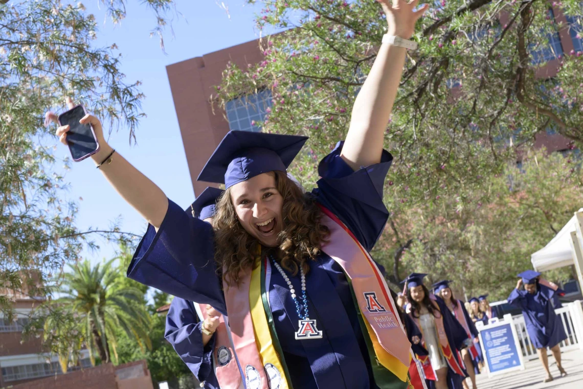 A young woman in a graduation cap and gown raises her arms in celebration as she leads a line of other graduates outside. 