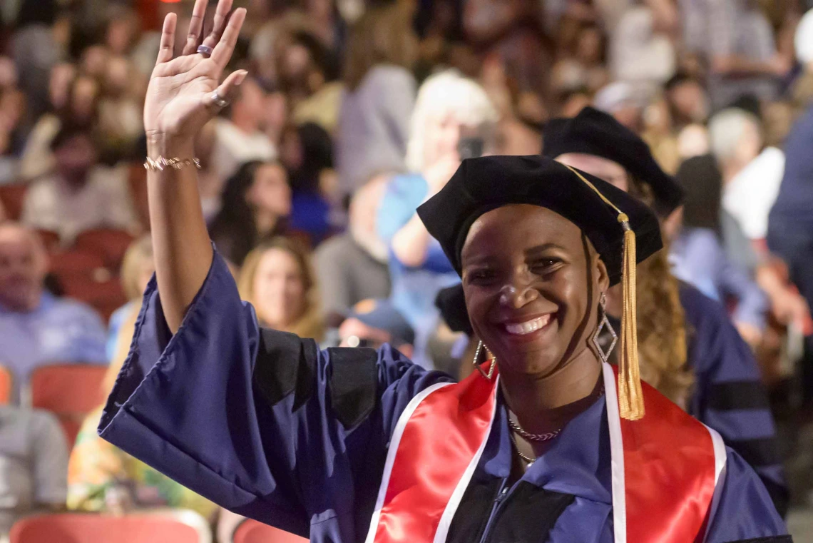 A woman wearing graduation regalia smiles and waves as she walks down an aisle of an auditorium filled with people. 