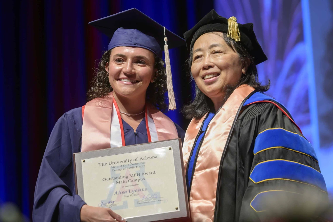 A young woman holding a certificate stands next to a professor onstage during a graduation ceremony. Both are wearing graduation regalia.