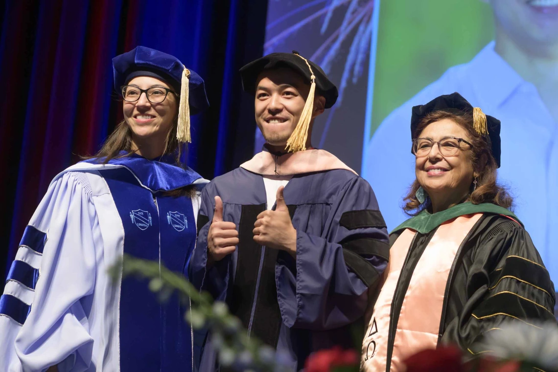 A young man gives two thumbs up as he stands between two professors. All are wearing graduation regalia on a stage and there is a projection of the man on a screen behind them. 