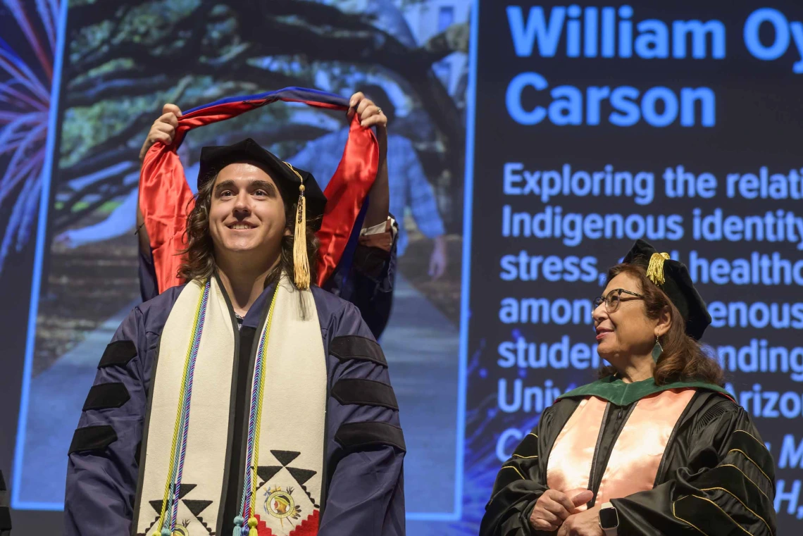 A young man wearing a graduation cap and gown smiles as a professor places a traditional graduation hood over his shoulders. Another professor looks on from the side. 