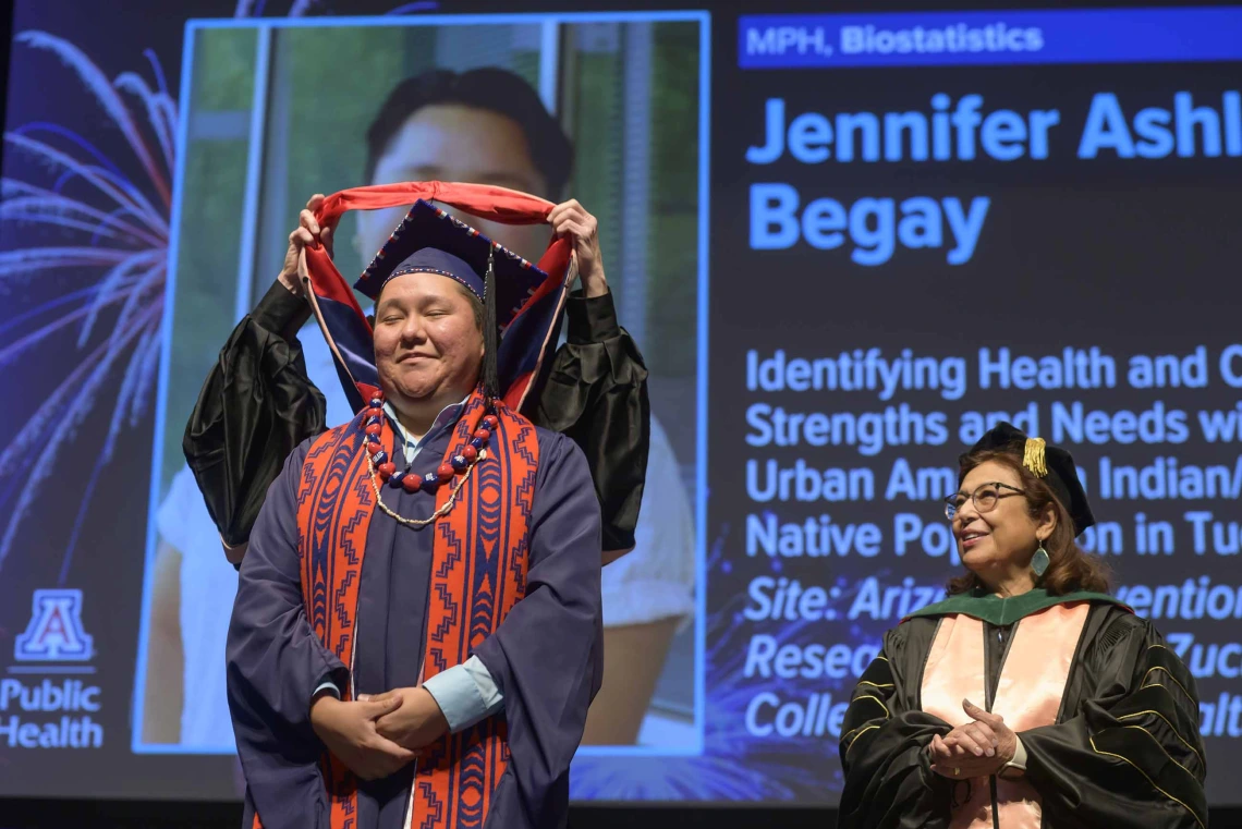 A young woman dressed in a graduation cap and gown closes her eyes and smiles as a professor places a graduation hood over her shoulders while another professor looks on.