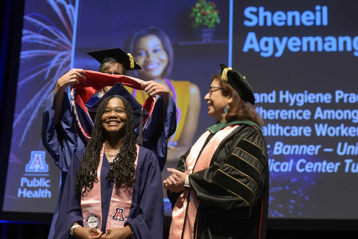 A young woman smiles as a professor places a ceremonial hood over her shoulders during a graduation ceremony while another professor looks on. All are wearing graduation caps and gowns. 