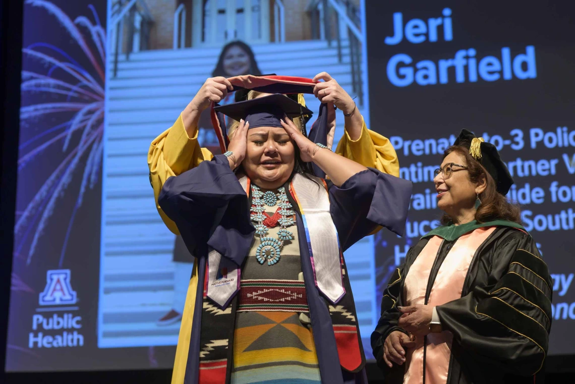 A young woman dressed in a graduation cap and gown and wearing a turquoise necklace and traditional Native American dress holds back tears as a professor places a graduation hood over her shoulders while another professor looks on.