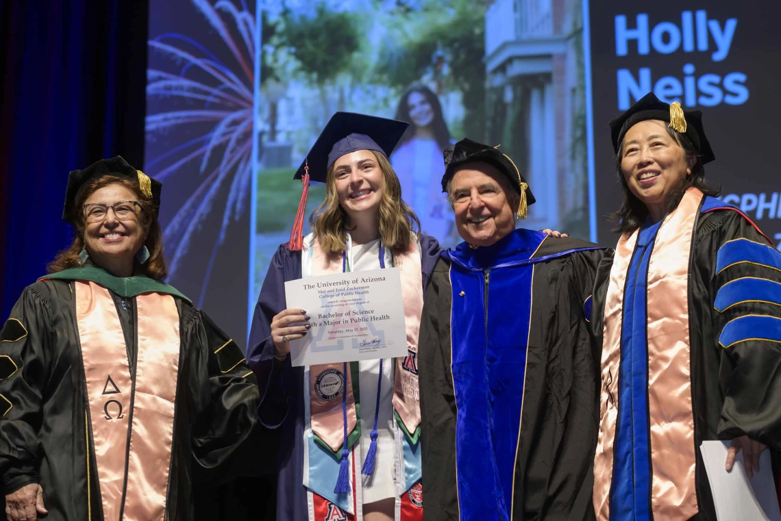 A young woman holds up a certificate and smiles while standing on a stage with several professors. All are wearing graduation regalia. 