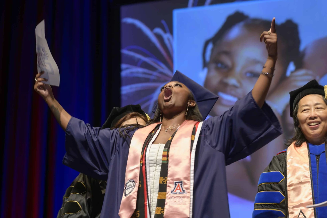 A young woman wearing a graduation cap and gown holds both of her arms up in the air in celebration during her graduation ceremony. 