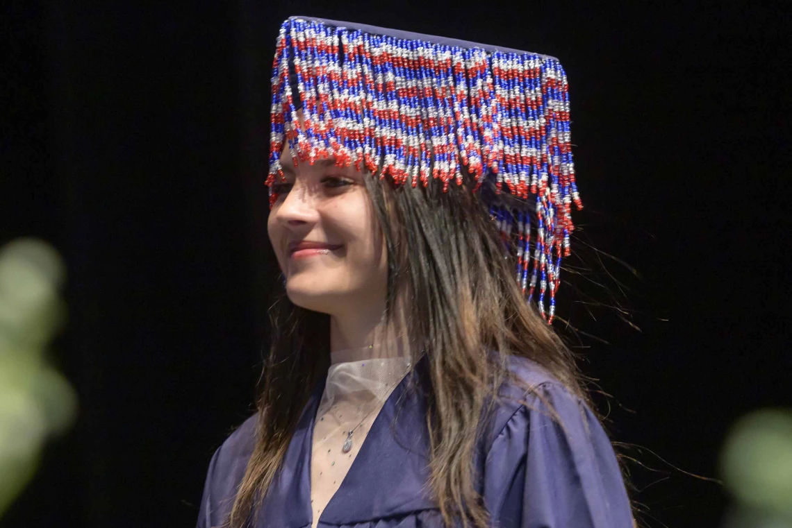 A young woman wearing a graduation gown and with beads hanging from her mortar board walks across a stage, smiling. 