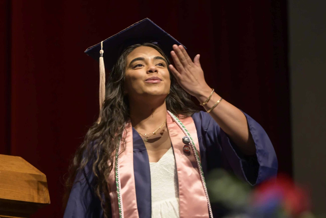 A young woman dressed in a graduation cap and gown blows a kiss as she walks across a stage during a graduation ceremony. 