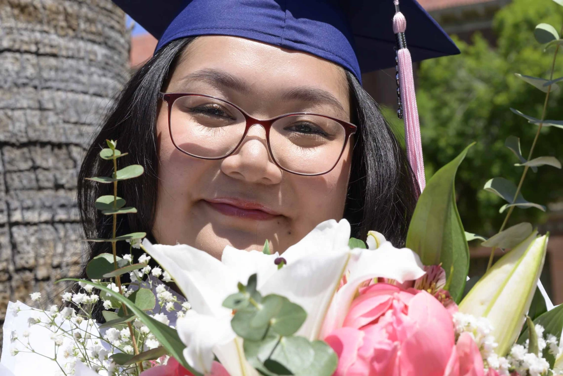 A young woman wearing a graduation cap smiles as she looks over a bouquet of flowers. 