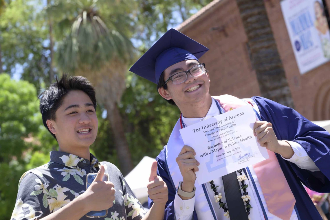 A young man wearing a graduation cap and gown smiles as he holds up his diploma while his friend looks on. 