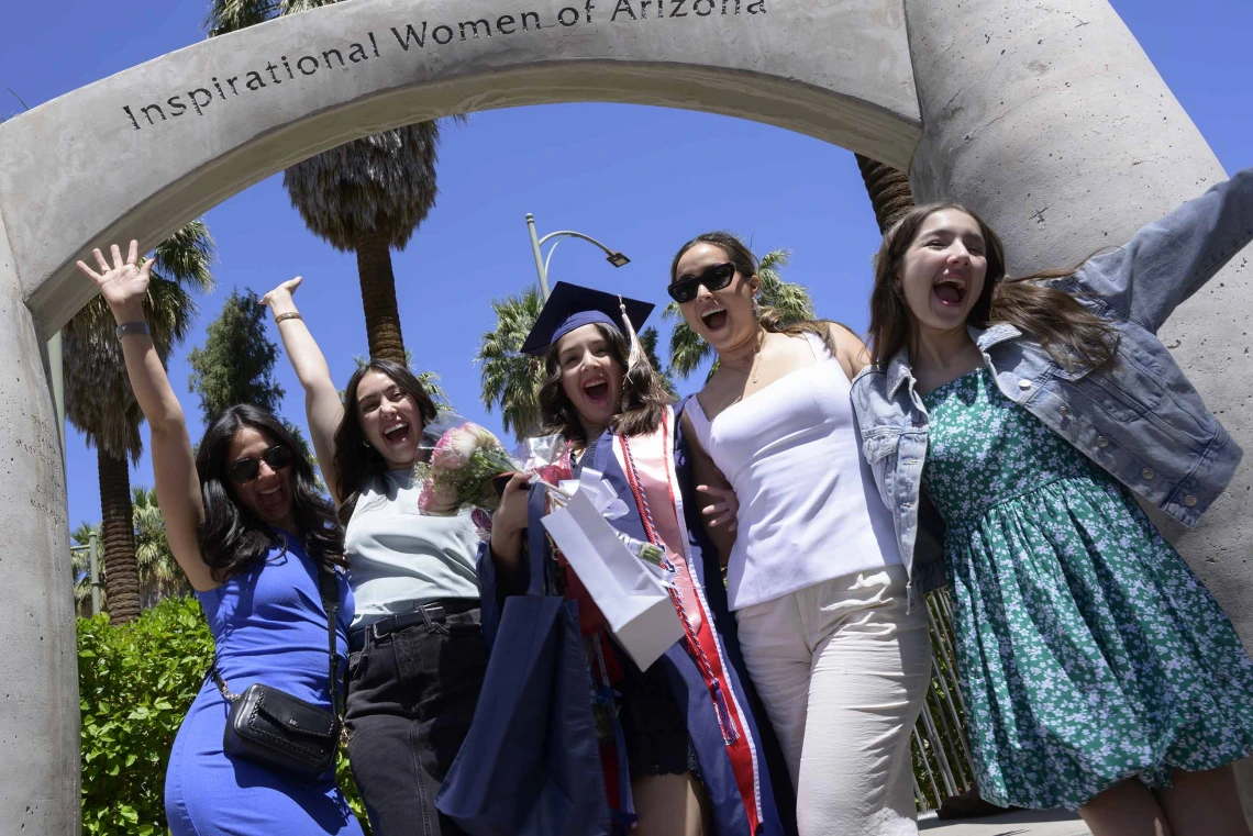 Five young women stand together, smiling and cheering, under a cement arch that reads “Inspirational Women of Arizona” outside. One of the women is wearing a graduation cap and gown. 