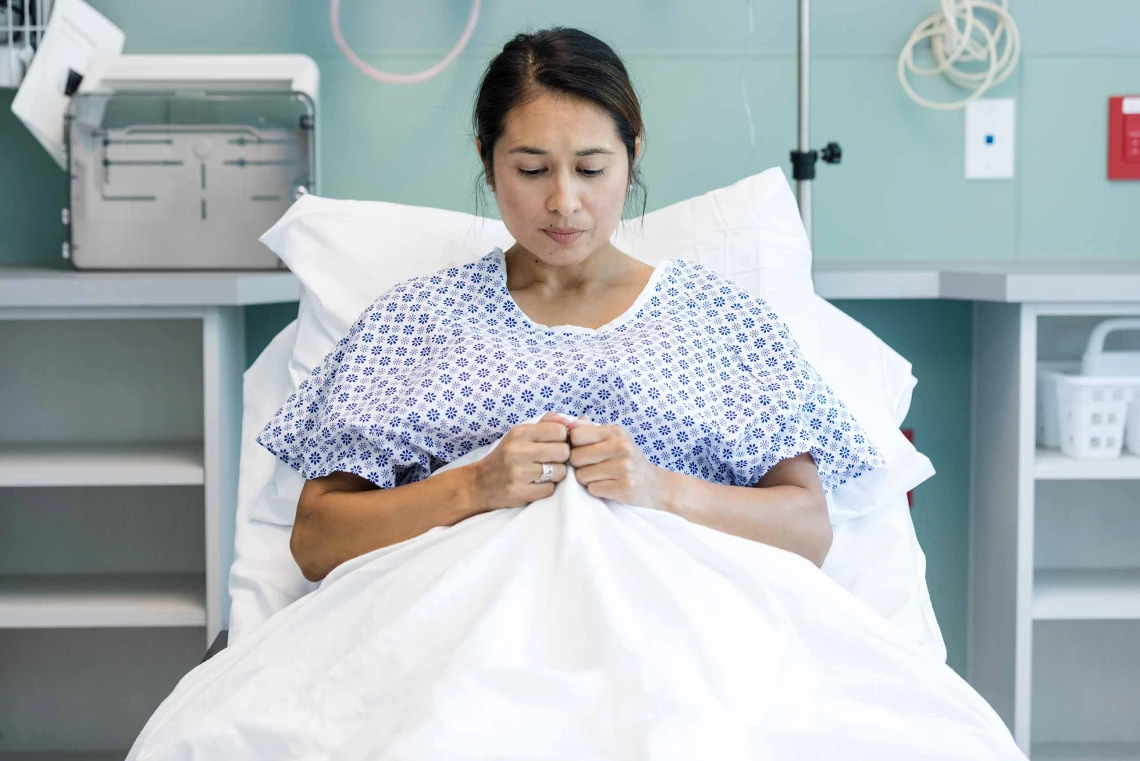 Woman wearing hospital gown laying on gurney with pensive look, clutching blanket with both hands