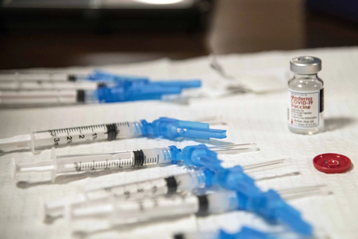 A table covered by a while cloth holding a vial with label “Moderna COVID-19 vaccine” and several syringes with needles ready for vaccination injections