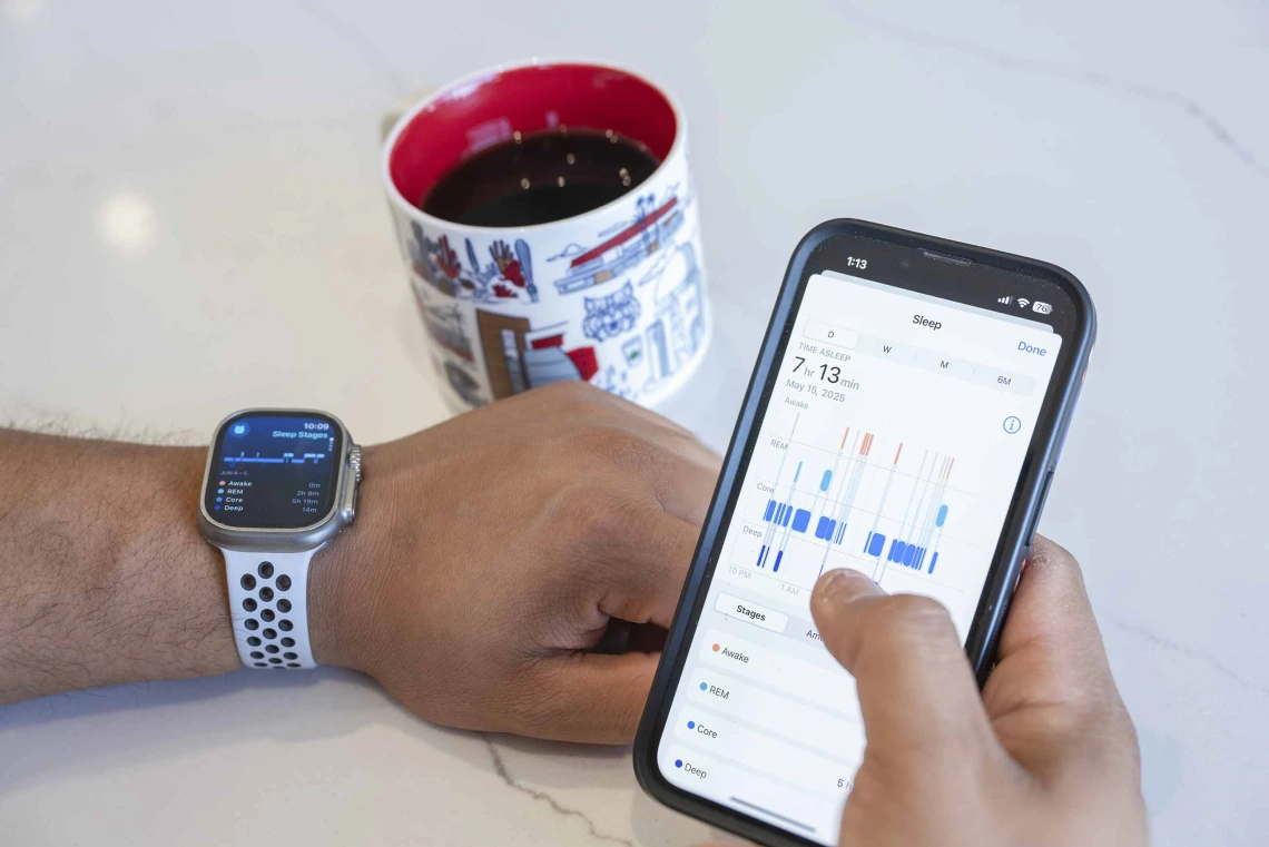 A person’s hands resting on a table, with an Apple Watch on the left wrist and right hand holding an iPhone displaying sleep date. A University of Arizona mug of coffee sits on the table