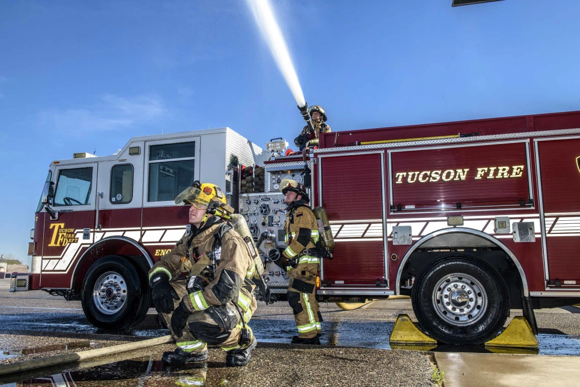 Two firefighters in the foreground and one spraying water from the top of a Tucson Fire firetruck